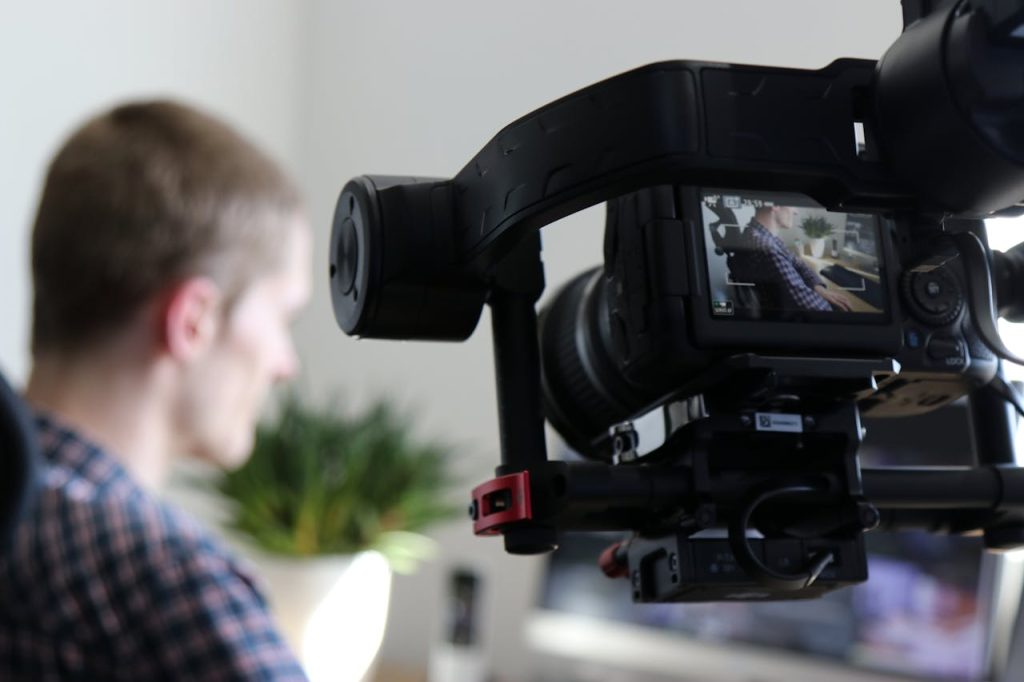 black-video-camera-turn-on-next-to-man-wears-black-and-gray-shirt-1046151 Close-up of camera filming a man in an indoor setting, highlighting professional video production